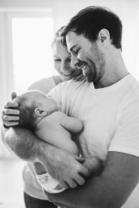 Ein berührendes Schwarz-Weiß-Porträt einer Familie, mit einer glücklichen Mutter und einem Vater, die ihr neugeborenes Baby liebevoll im Arm halten, während sie gemeinsam lachen.