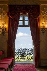 Ein Foto von innen des Traumzimmers im Schloss Burgeln in Schliengen. Im Fokus steht das romantische Fenster mit einer beeindruckenden roten Samtvorhang, einem Balkon und Blick auf die Schlossaußenanlagen an einem schönen sonnigen Tag. Aufgenommen von der Fotografin Isabela Campos.