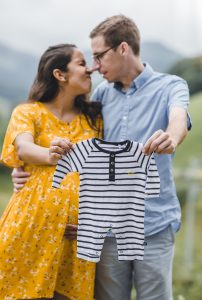 Familienfotoshooting - Mama und Papa halten ein niedliches Baby-Outfit vor der Kamera